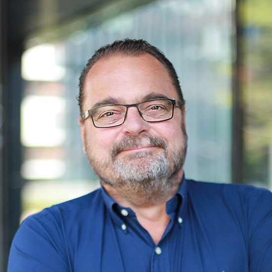 Natural Pharmaceuticals Movisan® Collagen II Portret uśmiechniętego mężczyzny w okularach i niebieskiej koszuli na rozmytym, nowoczesnym tle.