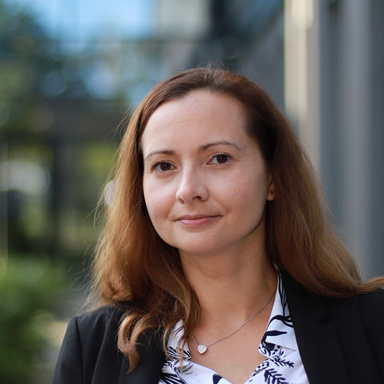 Natural Pharmaceuticals Movisan® Collagen II Portret uśmiechniętej kobiety o jasnobrązowych włosach w eleganckim ubraniu na tle miejskiej scenerii.