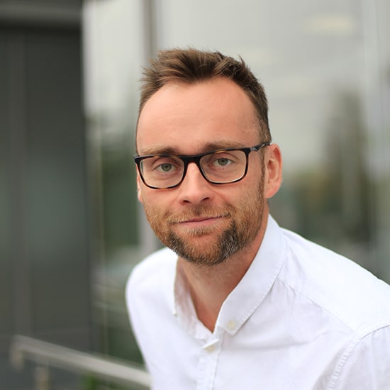 Natural Pharmaceuticals Movisan® Collagen II Portret mężczyzny w białej koszuli i okularach na tle nowoczesnego budynku, uśmiechnięty i pewny siebie.