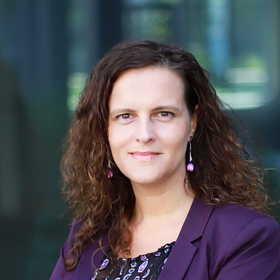 Natural Pharmaceuticals Movisan® Collagen II Portret kobiety o kręconych włosach w fioletowej marynarce na rozmytym tle biurowym.