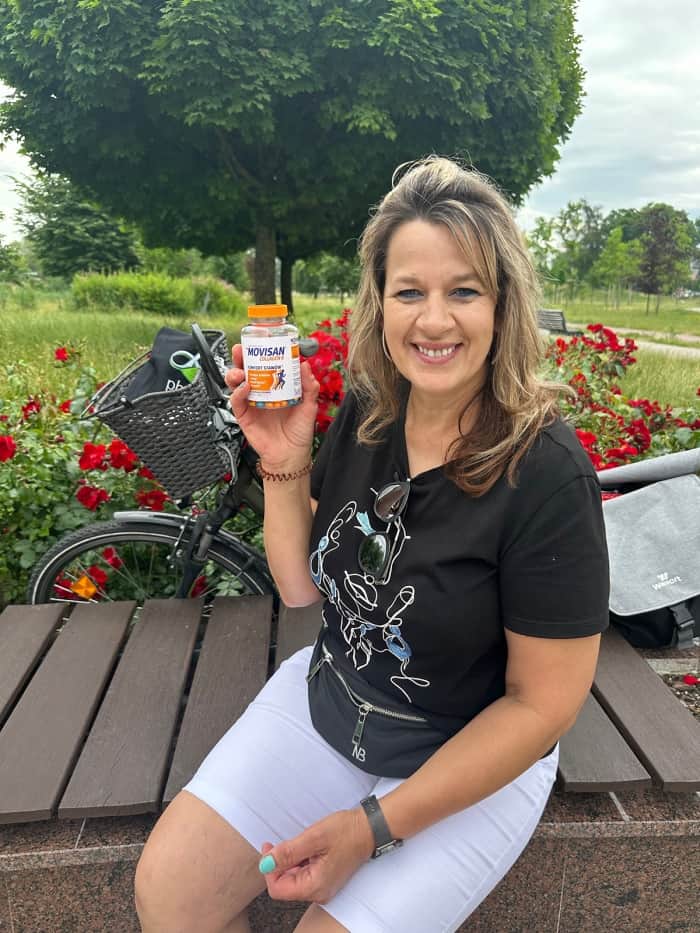 Natural Pharmaceuticals Movisan® Collagen II Kobieta siedzi na ławce w parku, trzymając butelkę suplementu Movisan na stawy, z rowerem i kwiatami w tle.