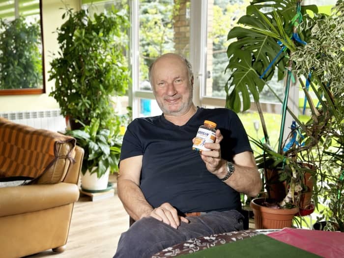 Natural Pharmaceuticals Movisan® Collagen II Mężczyzna siedzi w jasnym pokoju z roślinami, trzymając opakowanie suplementu diety w dłoni.