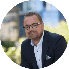 Natural Pharmaceuticals Movisan® Collagen II Portret uśmiechniętego mężczyzny w okularach i marynarce na rozmytym miejskim tle.